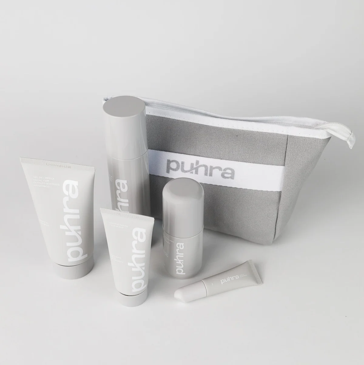 Quatro mãos segurando produtos da linha Puhra: dois tubos, um frasco spray e um sérum, todos em embalagens minimalistas na cor bege/cinza claro